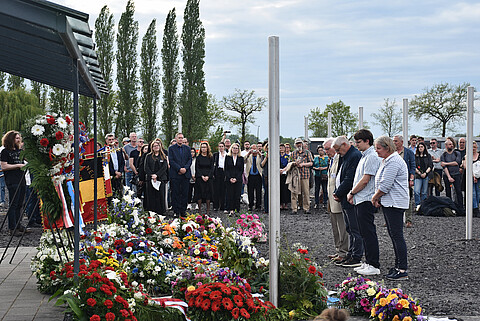 Kranzzeremonie am ehemaligen Arrestbunker mit vielen Blumengestecken, davor steht eine Gruppe von vier Personen, im Hintergrund weitere Personen.