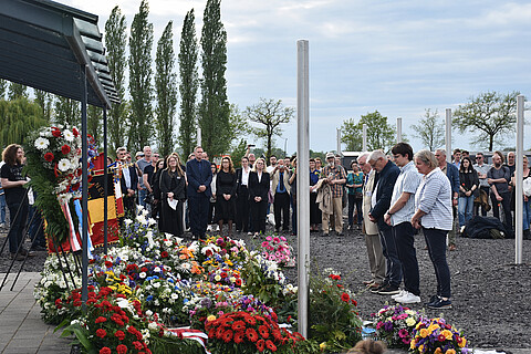 Kranzzeremonie am ehemaligen Arrestbunker mit vielen Blumengestecken, davor steht eine Gruppe von vier Personen, im Hintergrund weitere Personen.