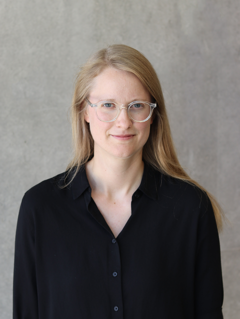 Anna trägt ein schwarzes Hemd und eine Brille und blickt lächelnd in die Kamera