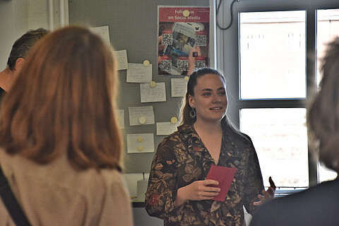 Uliana bei der Vorstellung ihrer Ausstellung. Sie hält einen roten Umschlag in der Hand. Eine junge Dame erzählt etwas, drei Personen hören zu und sind von hinten zu sehen.