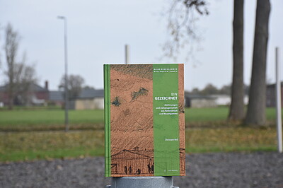 Buch mit Zeichnung und grünen grafischen Elementen, im Hintergrund unscharf Wiese und Bäume