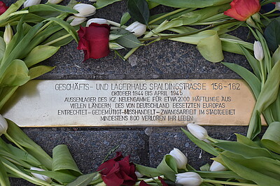 Auf der rechteckigen Messingplatte steht: „Geschäfts- und Lagerhaus Spaldingstraße 156-162. Oktober 1944 bis April 1945. Außenlager des KZ Neuengamme für etwa 2000 Häftlinge aus vielen Ländern des von Deutschland besetzten Europas. Entrechtet – gedemütigt – misshandelt Zwangsarbeit im Stadtgebiet. Mindestens 800 verloren ihr Leben“. Um die Stolperschwelle sind Tulpen verteilt.
