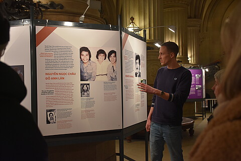 Ein Mann deutet auf einer Ausstellungstafel auf die Schrift und Fotos gedruckt sind.