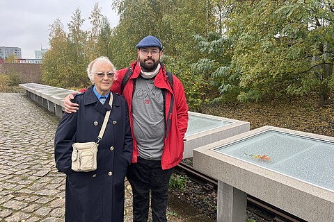 : Eine ältere Frau und ein Mann stehen vor einer Reihe Namenstafeln. Der Man hat seinen Arm um die Schultern der älteren Frau gelegt. Hinter den Namenstafeln sind Bahngleise