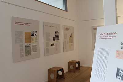 Tafeln mit Text und Fotos, die an der Wand hängen, davor Hocker.