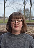 Clara Mansfeld trägt eine Brille und ein graues Kleid, im Hintergrund Bäume und die angedeuteteten Fundamente einer ehemaligen Baracke