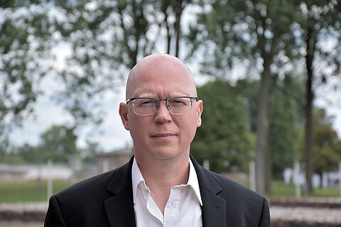 Karsten Uhl trägt eine Brille, ein weißes Hemd und schwarzes Jacket. Im Hintergrund Bäume und die Fundamente ehmaliger Baracken.