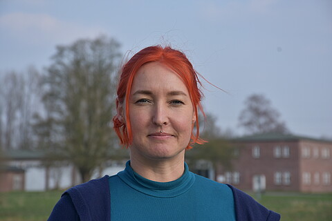 Gisela hat rote kinnlange Haare und ein Medusa-Piercing, im Hintergrund Gebäude und Bäume.