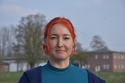 Gisela hat rote kinnlange Haare und ein Medusa-Piercing, im Hintergrund Gebäude und Bäume.