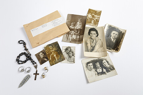 Bild von Familienfotos, Schmuck und einem Kreuz neben einer Papiertasche.  Dies sind Effekten von Neonella Doboitschina, die u.a. in einem Außenlager des KZ Neuengamme inhaftiert war