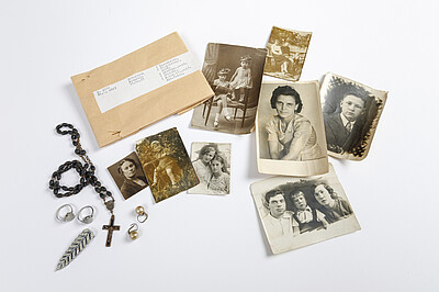 Bild von Familienfotos, Schmuck und einem Kreuz neben einer Papiertasche.  Dies sind Effekten von Neonella Doboitschina, die u.a. in einem Außenlager des KZ Neuengamme inhaftiert war