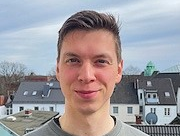 Portraitfoto eines jungen Mannes im grauen Sweatshirt vor Häusern im Hintergrund